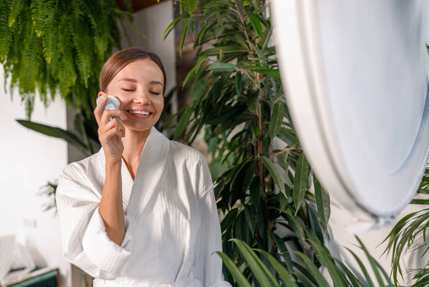 Glückliche junge Frau im Bademantel lächelt und genießt die Verwendung von Silikon-Gesichtsbürste zum Peeling der Haut, steht im Badezimmer mit grünen Pflanzen dekoriert - Foto, Bild