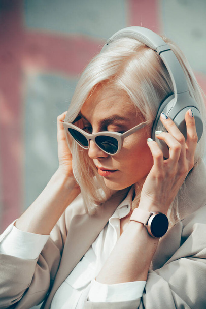 Πορτρέτο της ξανθιάς γυναίκας με ακουστικά που ακούει μουσική με γυαλιά ηλίου. Hipster μοντέλο μόδας φορούν μοντέρνα ασύρματα ακουστικά απολαμβάνουν να ακούσετε νέα δροσερή μουσική. - Φωτογραφία, εικόνα