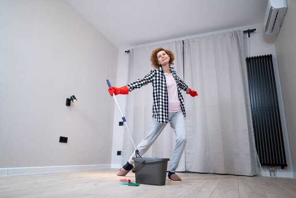 Ενθουσιασμένη ώριμη γυναίκα που απολαμβάνει το καθαριστήριο, χορεύει ενώ πλένει το πάτωμα. Ευτυχισμένη ηλικιωμένη γυναίκα που απολαμβάνει να καθαρίζει πατώματα πριν μετακομίσει σε νέο διαμέρισμα. Έννοια της οικιακής εργασίας και της οικιακής οικονομίας. - Φωτογραφία, εικόνα