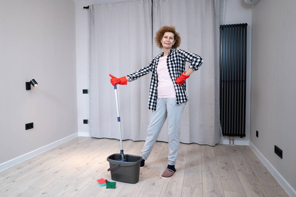 Pessoas, tarefas domésticas e conceito de limpeza. Mulher sênior feliz em luvas de proteção limpa o chão e dança em casa em apartamento vazio antes de se mudar para uma nova casa. Mulher excitada desfrutando de limpeza. - Foto, Imagem