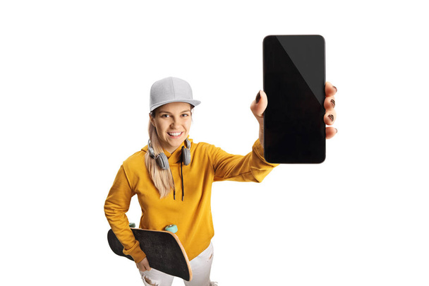Young female with headphones and a skateboard holding a smartphone in front of camera isolated on white background - Φωτογραφία, εικόνα
