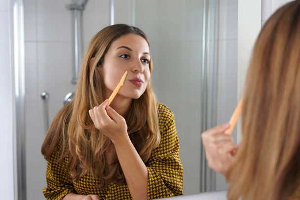 Depilación facial. Hermosa joven afeitándose la cara con una navaja en casa. Mujer bonita usando afeitadora en el baño. - Foto, imagen