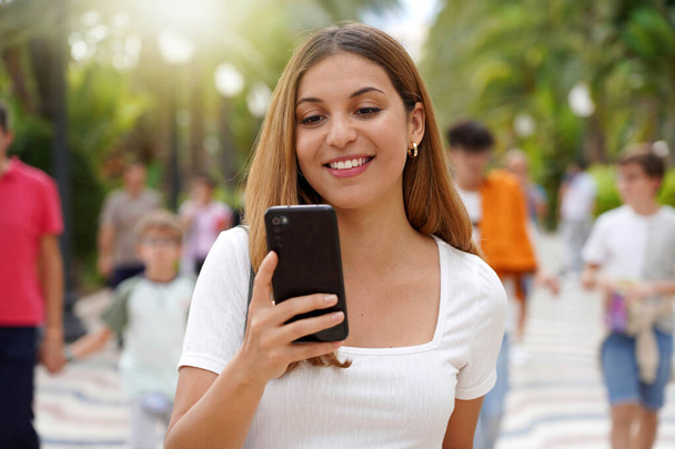 Fröhliche, fröhliche junge Frau schaut auf ihr Smartphone, wenn sie mit verschwommenen Menschen im Hintergrund auf der Straße läuft. Millennial Mädchen mit Handy-App im Freien. Lifestyletechnologiekonzept für Teenager. - Foto, Bild