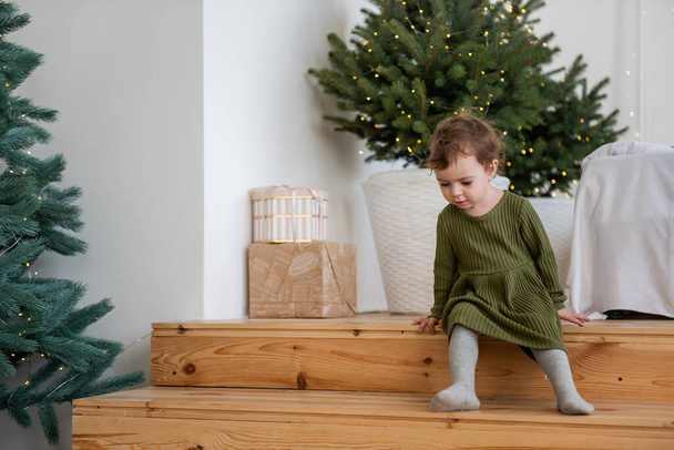 Niña en vestido verde cálido se sienta en los escalones de madera junto al árbol de Navidad con guirnalda en la sala de estar loft blanco. Minimalista estilo escandinavo del apartamento. Estilo de vida festivo decorado - Foto, Imagen