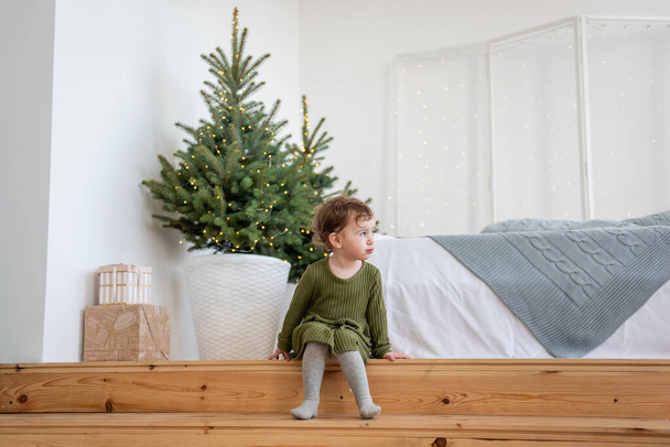Meleg zöld ruhás kislány ül a fa lépcsőn a karácsonyfa mellett, koszorúval a fehér tetőtéri nappaliban. Minimalista skandináv stílus a lakás. Ünnepi díszítésű életmód - Fotó, kép