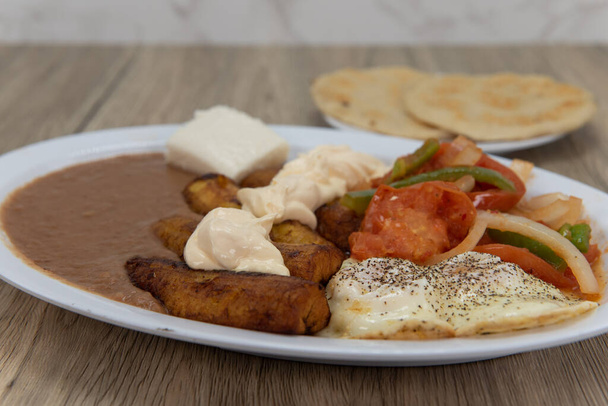 Plantains frits dans une assiette avec haricots frits, légumes frits et œufs servis côté ensoleillé avec du pain pupusa pour un repas latino. - Photo, image