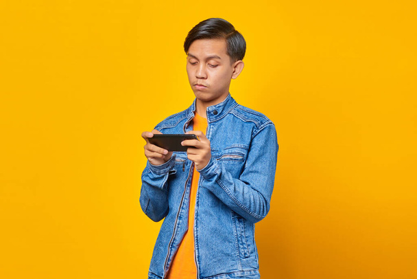 азиатский человек, использующий мобильный телефон играть в игру с сердитым лицом на желтом фоне - Фото, изображение
