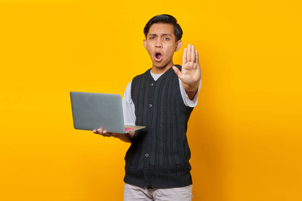 Young Asian man using laptop and  making stop gesture with her hands on yellow background - Photo, Image