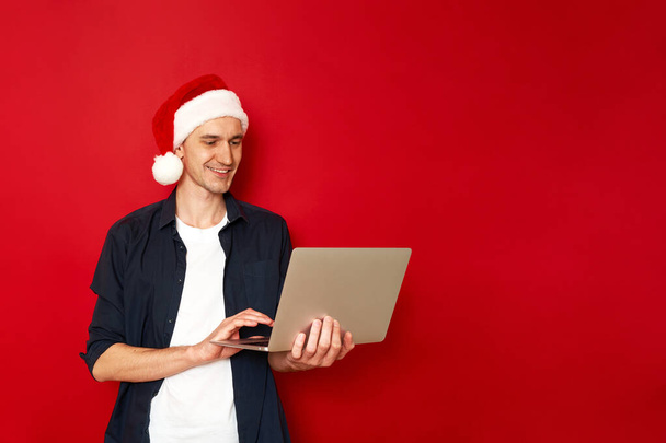Smějící se mladý muž v ležérní modré košili vánoční klobouk pózovat izolované na červeném pozadí studiového portrétu. Lidé upřímné emoce životní styl koncept. Zesměšnit kopírovací prostor. Práce na přenosném počítači - Fotografie, Obrázek
