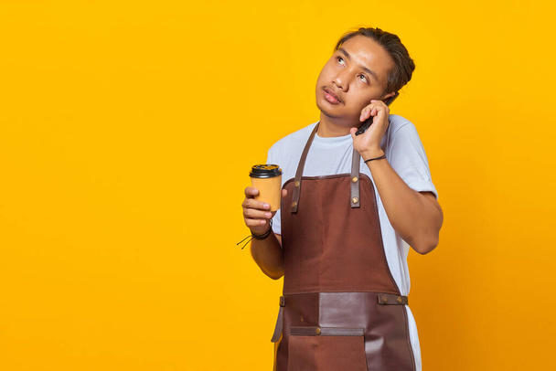 Portret atrakcyjnego Azjaty rozmawiającego na smartfonie i trzymającego filiżankę kawy na żółtym tle - Zdjęcie, obraz