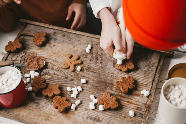 Children decorate the Christmas gingerbread man with white frosting on a wooden background. Christmas sweets with cocoa and marshmallows for children. Winter vacation at home. Fun time in the kitchen. - Photo, image