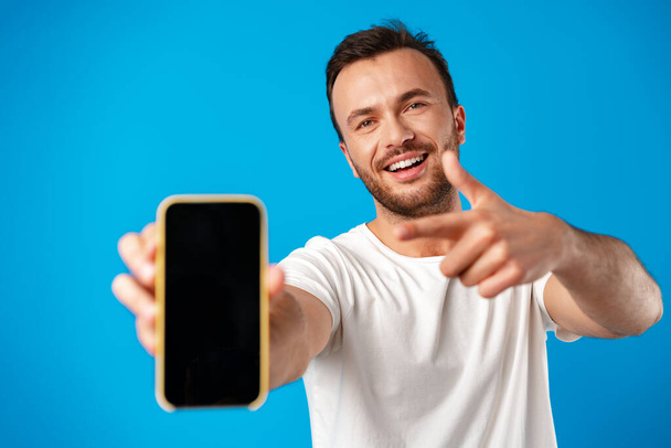 Portrait of young man advertising new smartphone, showing it to camera against blue background - Fotografie, Obrázek
