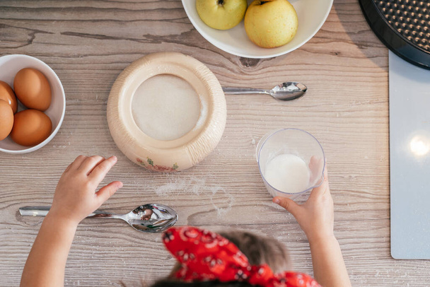 Mains de petite fille dans le bandeau rouge cuire tarte aux pommes. L'enfant met du sucre dans une tasse à mesurer. Un gamin qui cuisine. Vue du dessus - Photo, image