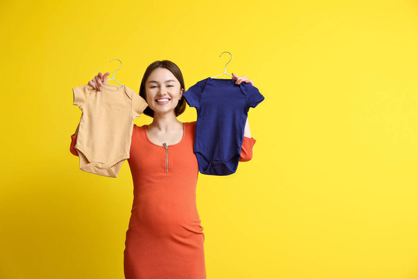 Νεαρή έγκυος γυναίκα με μωρουδιακά κοστούμια σε κίτρινο φόντο - Φωτογραφία, εικόνα