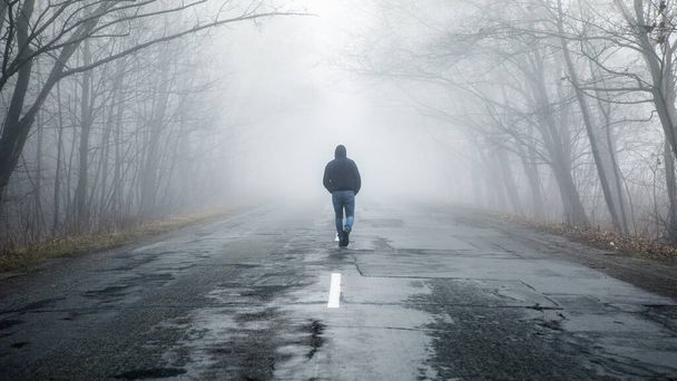 Eenzame man loopt in de mist weg. Landelijk landschap met weg in de ochtendmist. Warme herfstkleuren. Donkere mysterieuze achtergrond - Foto, afbeelding