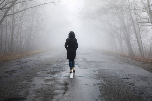 Mujer solitaria caminando en el camino de la niebla. Paisaje rural con carretera en niebla matutina. Colores cálidos de otoño. Fondo oscuro misterioso - Foto, imagen
