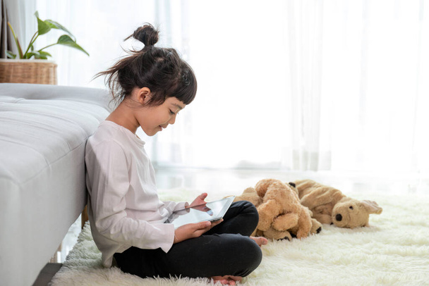 Mała dziewczynka leżąca na podłodze w salonie z tabletem patrząc na ekran urządzenia oglądania kreskówek cieszyć słuchanie audiobook, przedszkola nowoczesny tech użytkownika kontroli rodzicielskiej bezpieczeństwa, gadżet nadużywania koncepcji - Zdjęcie, obraz