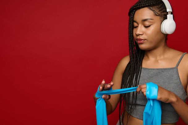 青い伸縮性のあるストラップを手にした無線ヘッドフォンを手にしたアフリカのスポーツ女性が、空間のコピーを手に赤い色の背景に立っている姿がクローズアップされている。 - 写真・画像