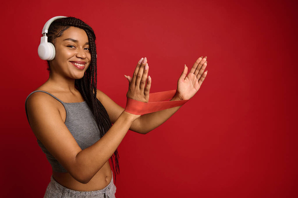 Mujer bonita de etnia africana con rastas elegantes que usan banda elástica de fitness roja mientras hacen ejercicios de estiramiento en los brazos, sonríe con una sonrisa dental, aislada en un fondo de color con espacio para copiar - Foto, Imagen