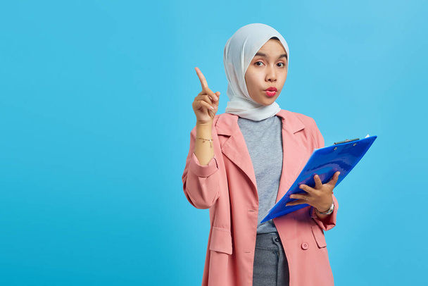 Portrait of a beautiful smiling woman holding a folder standing and pointing up on a blue background - Foto, Imagem