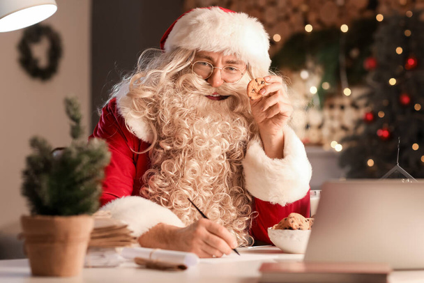 Santa Claus eating tasty cookies at home in evening - Фото, изображение