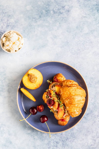 Fresh croissant with cream cheese, nectarine and cherry on blue ceramic plate on a blue background. Breakfast with croissant and juice. - Photo, Image