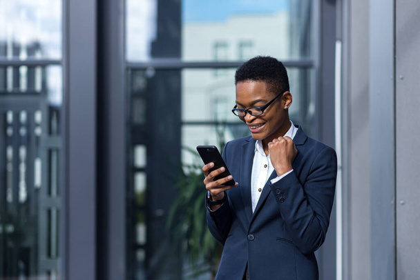 Happy and successful business woman rejoices in winning and happy day, celebrates, african american smiling holds phone in hands - Фото, зображення