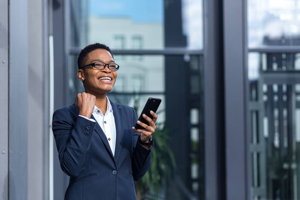 Happy and successful business woman rejoices in winning and happy day, celebrates, african american smiling holds phone in hands - 写真・画像