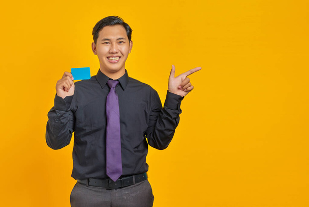 Smiling asian businessman holding a credit card while pointing finger at copy space on yellow - Photo, Image