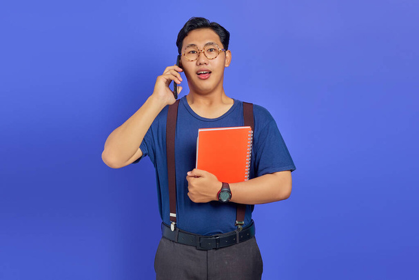 Shocked young student talking on smartphone while holding notebook on purple background - Φωτογραφία, εικόνα