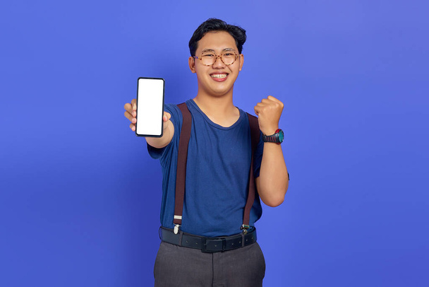 Asian young man showing blank smartphone screen with hands to camera on purple background - Photo, Image