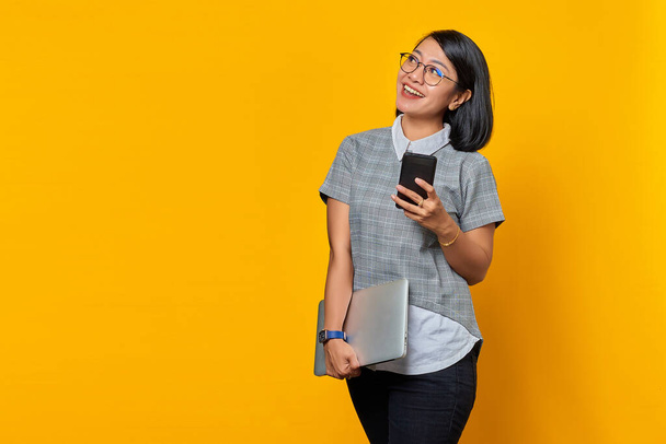 Cheerful beautiful young woman looking at mobile phone screen and holding laptop on yellow background - Фото, изображение