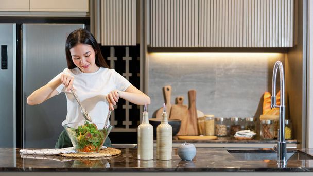 Salatkonzept steht die Frau mit weißem T-Shirt und dunkelgrüner Hose an der Küche und kocht eine Schüssel grünen Salat. - Foto, Bild