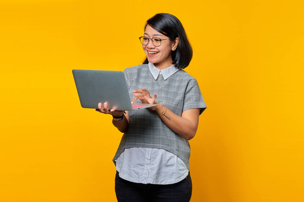 Веселая молодая азиатка держит и использует ноутбук изолирован на желтом фоне - Фото, изображение