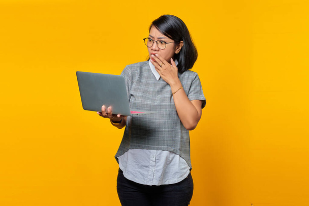 Asiatin mit Brille überrascht beim Öffnen eingehender E-Mails auf Laptop isoliert auf gelbem Hintergrund - Foto, Bild