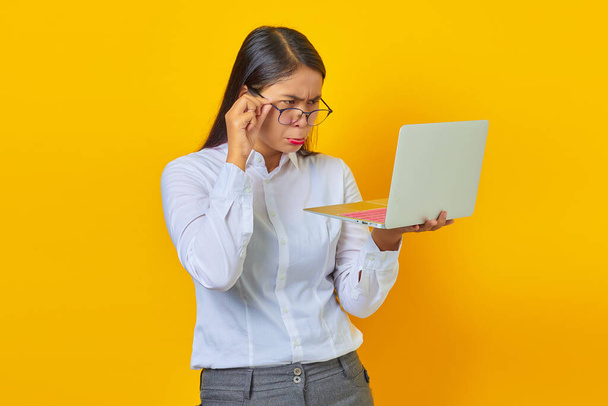 Verrast jonge zakenvrouw op zoek naar laptop en het houden van glazen op gele achtergrond - Foto, afbeelding