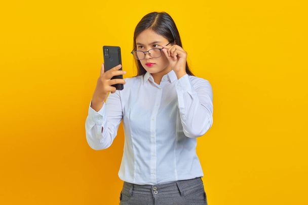 Wütende junge Asiatin benutzt Handy und nimmt Brille isoliert über gelbem Hintergrund - Foto, Bild