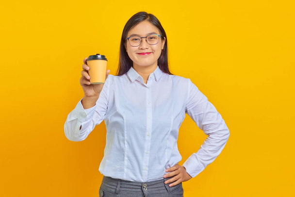 Photo of smiling and cheerful Asian business woman wearing white shirt holding freshly bought cup of coffee on yellow background - Photo, Image