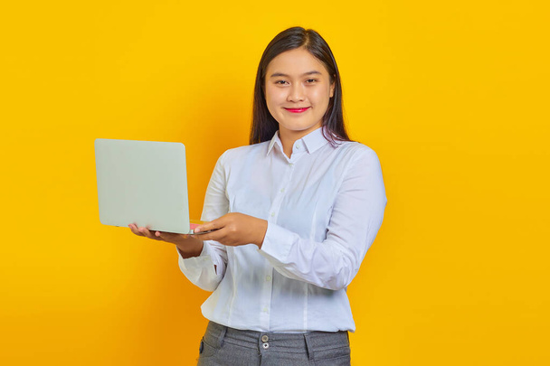 Zdjęcie wesołej młodej kobiety biznesu z pewną siebie twarzą za pomocą laptopa i patrząc na aparat odizolowany na żółtym tle - Zdjęcie, obraz