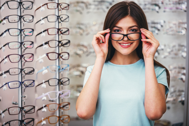 Young woman is choosing a glasses in optician store. - Фото, изображение