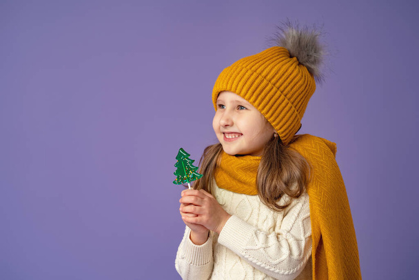 Little European girl of 5 years old with blonde hair in a yellow hat and scarf, holding a candy in the form of a green Christmas tree in her hand, stands smiling happily on a purple background. - Photo, image