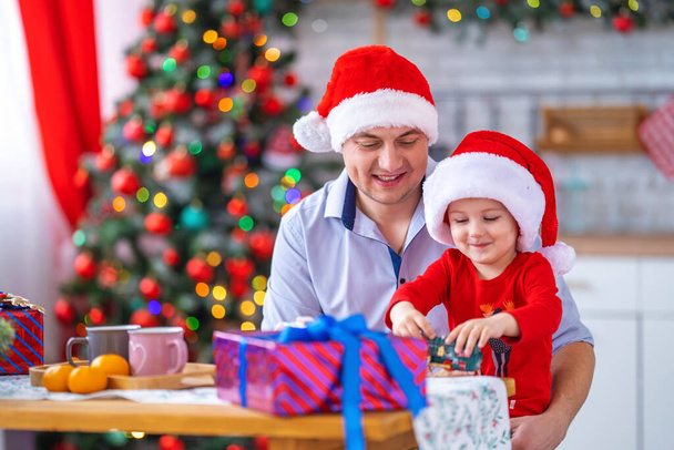 Merry Christmas and happy holidays. Loving dad with cheerful son in Santa hats, playing at table in kitchen on Christmas morning. Happy family, parent and child have fun opening gifts and playing - Φωτογραφία, εικόνα