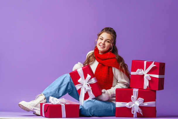 attractive beautiful girl with curly hair in white sweater and red scarf, sitting with gift boxes on blue background in studio. teenager smiles happily and hugs red gift box in mittens. Advertising. - Foto, Bild
