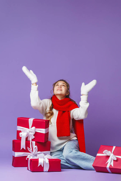 attractive beautiful girl with curly hair in white sweater and red scarf, sitting with gift boxes on blue background in studio. teenager smiles happily and raises his mittened hands up. Advertising. - Фото, изображение
