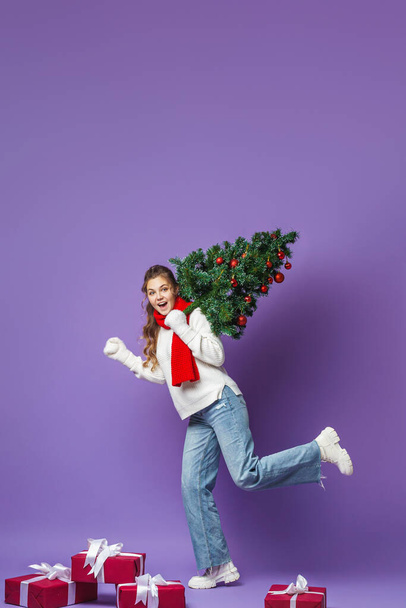 happy teenage girl with curly hair in a knitted sweater and a red scarf, full-length, with Christmas tree decorated with red Christmas balls on her shoulder, walks on purple background. Dynamic image. - Photo, image