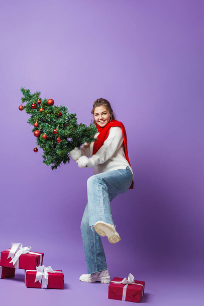 happy teenage girl with curly hair in a knitted sweater and a red scarf, full-length, with Christmas tree decorated with red Christmas balls on her shoulder, walks on purple background. Dynamic image. - Foto, Bild