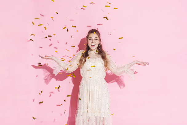 Merry Christmas. A beautiful attractive girl with wavy hair in a light shiny dress dancing under golden falling, flying confetti on a pink background in the studio, filmed with harsh light. Copy space - Foto, Imagen