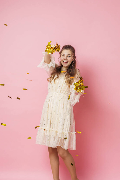 Merry Christmas. A beautiful attractive girl with wavy hair in a light shiny dress dances under golden falling, flying confetti on a pink background in the studio. In full growth. Copy space - Foto, immagini