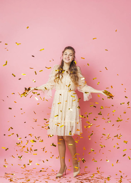 Merry Christmas. A beautiful attractive girl with wavy hair in a light shiny dress dances under golden falling, flying confetti on a pink background in the studio. In full growth. Copy space - 写真・画像