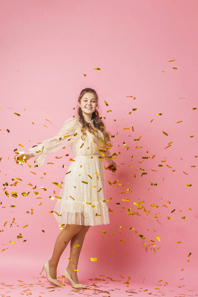 Merry Christmas. A beautiful attractive girl with wavy hair in a light shiny dress dances under golden falling, flying confetti on a pink background in the studio. In full growth. Copy space - Foto, Imagen